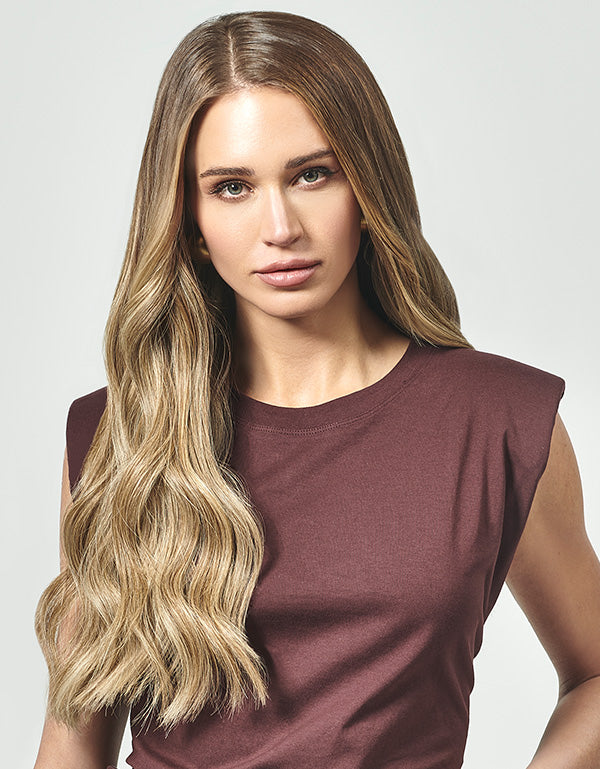 Model with long, wavy blonde hair, wearing a sleeveless brown top, posing against a neutral background.