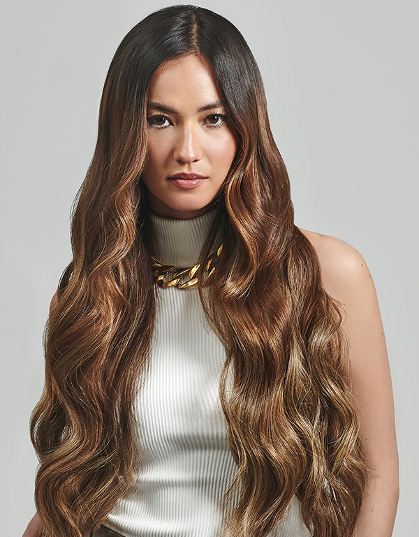 A woman with long wavy brown weft hair extensions wearing a white top, smiling softly.